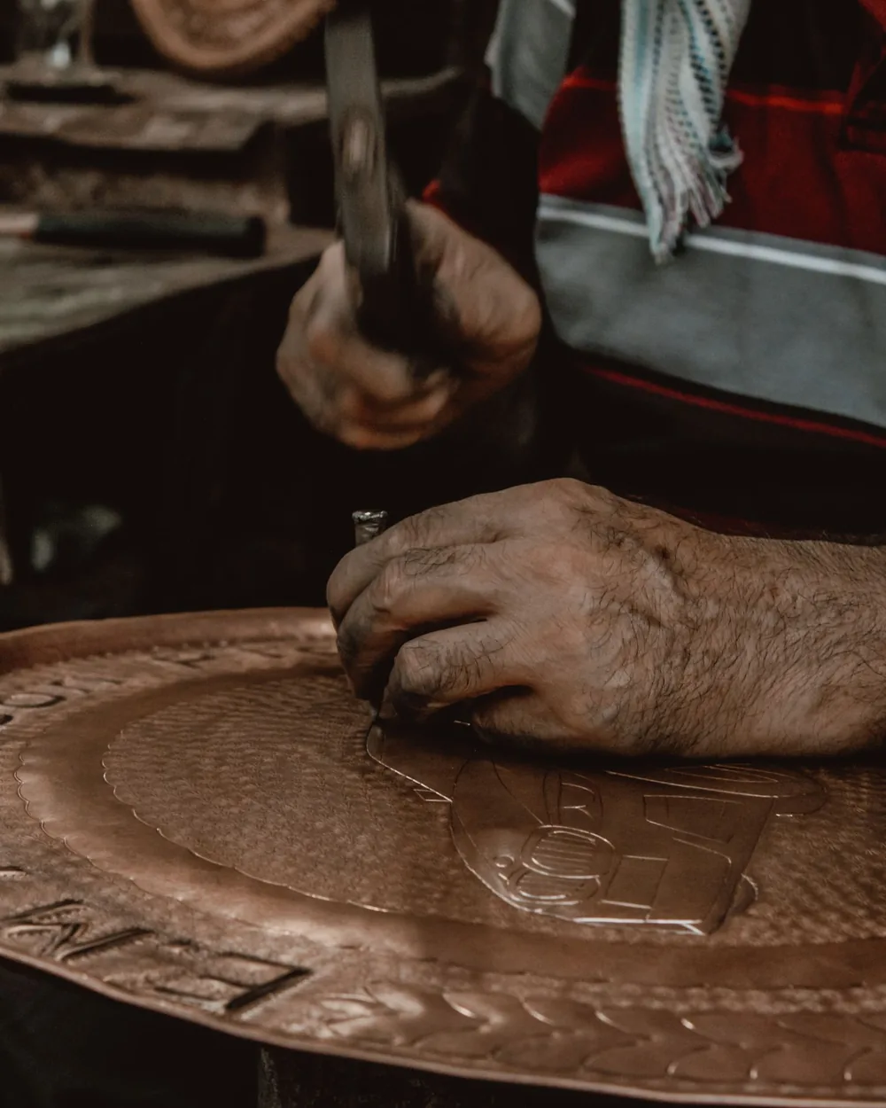 Manos sosteniendo un cincel sobre una bandeja de cobre mientras el martillo golpea el mango y marca el trazo en el metal.